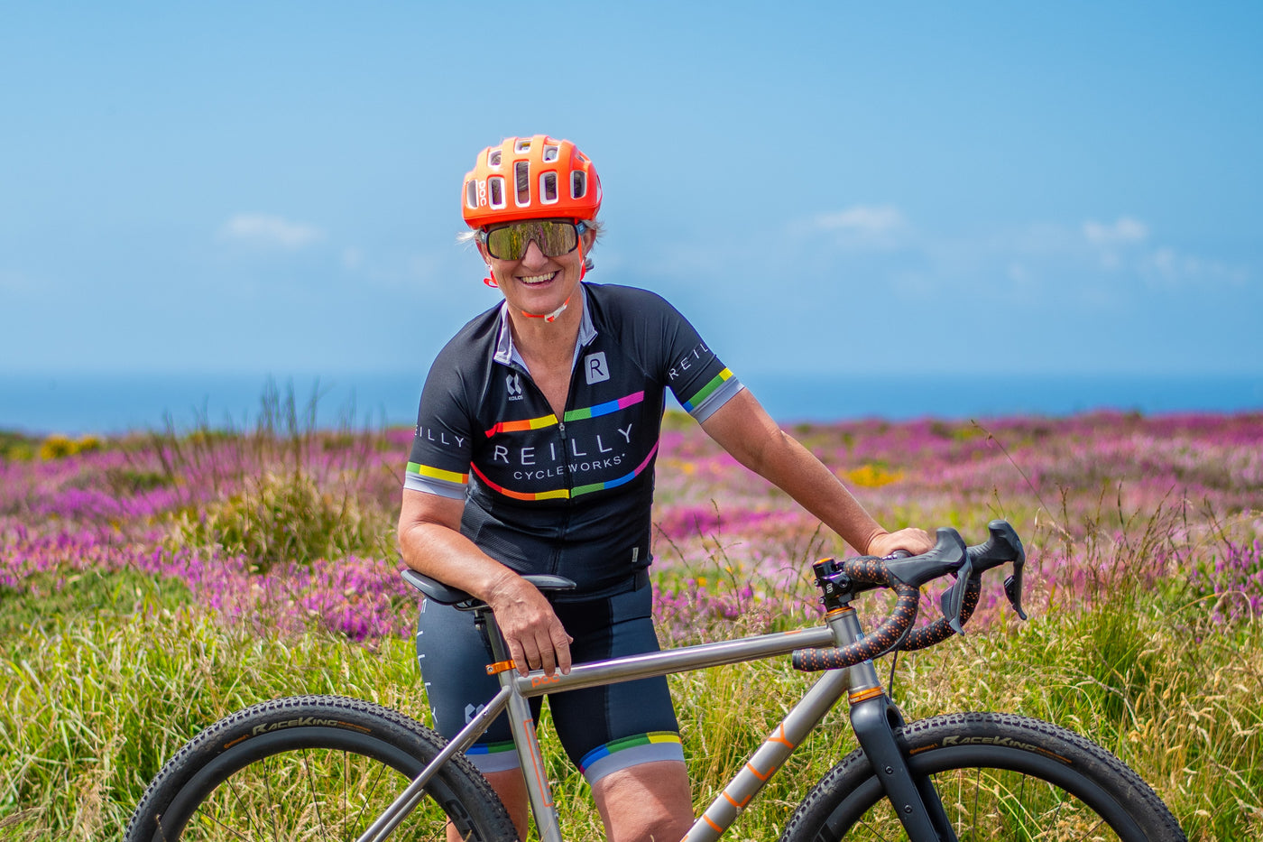 Smiling woman with orange cycle helmet stands behind her bike in a field of heather.