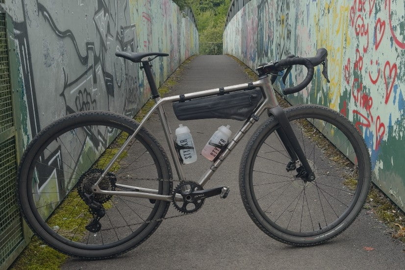 A stunning titanium gravel bike stands on a railway bridge with graffiti walls.