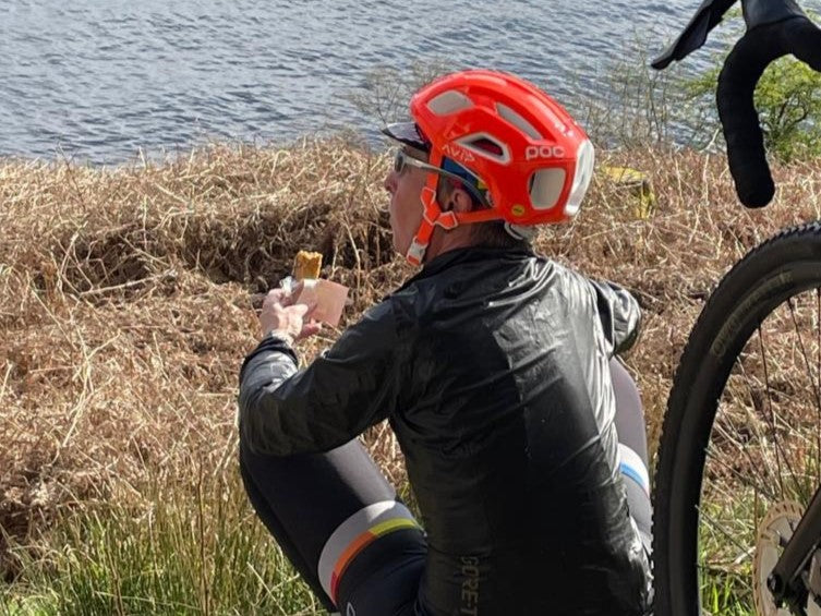 Woman in orange cycle helmet eats a snack looking out to to a lake.