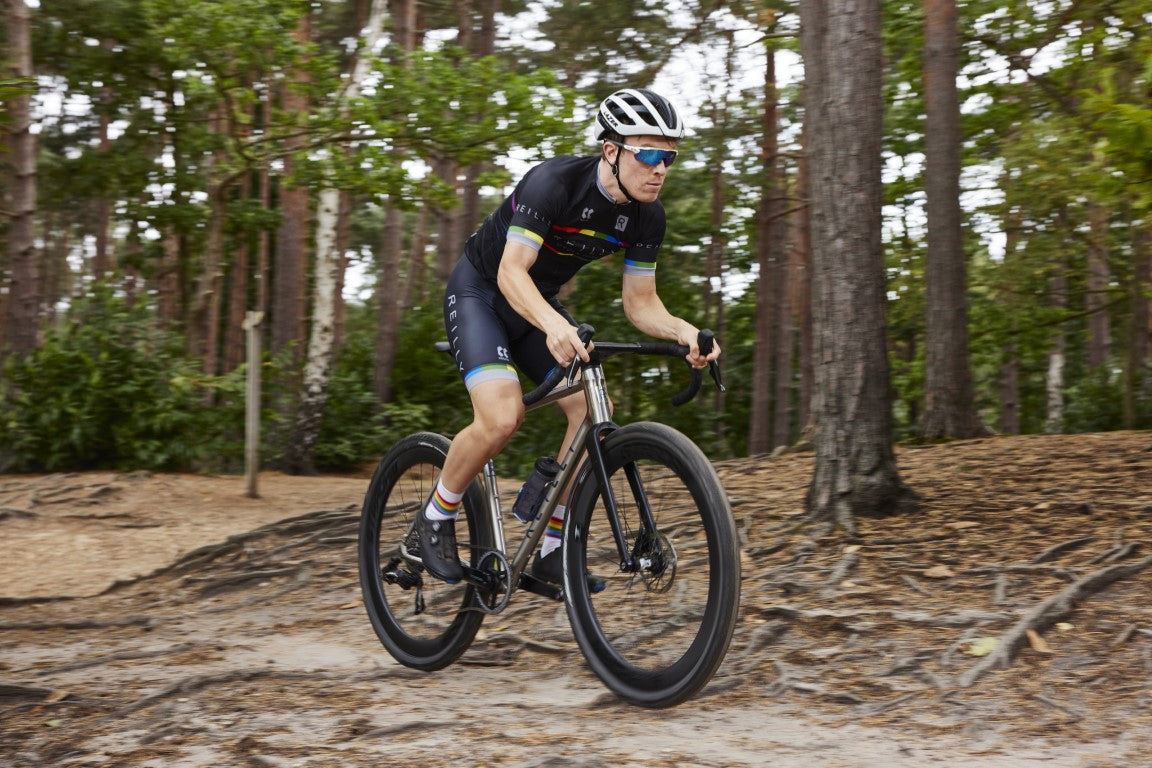 A serious looking man cycles a titanium gravel race bike through a forest.