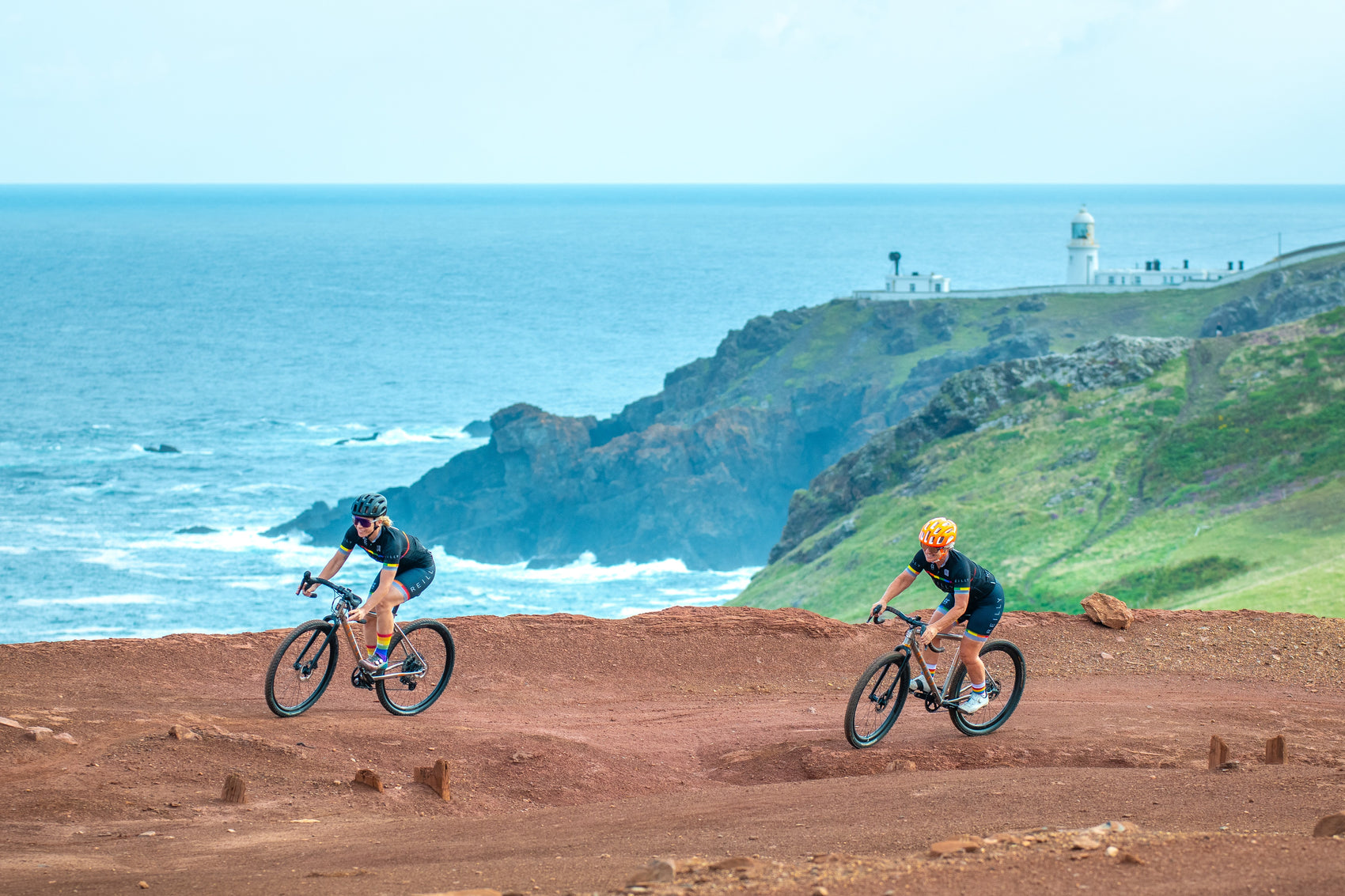 Two women on titanium gravel bicycles cycle on an earth path with cliffs and sea in the background.