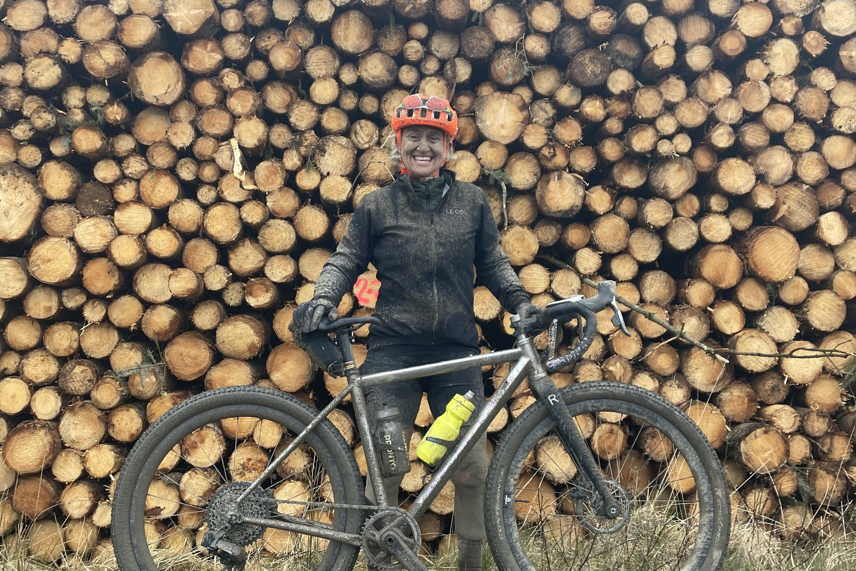 A muddy woman stands with her bike in front of a log pile with Dirty Reiver logo.