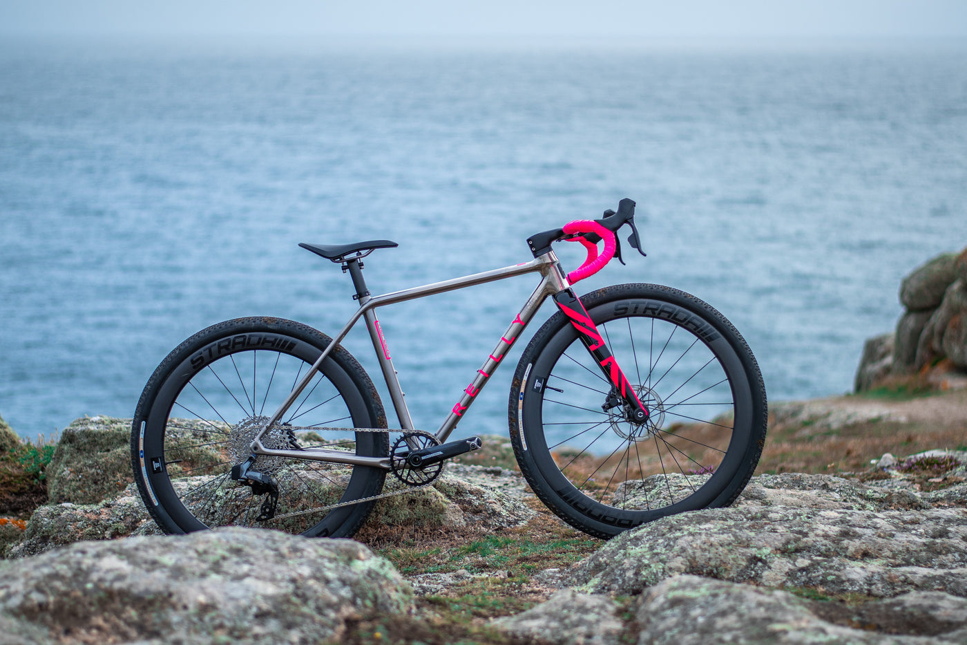 A titanium gravel bike with striking pink fork and bars on a rocky ground with the sea in the background.