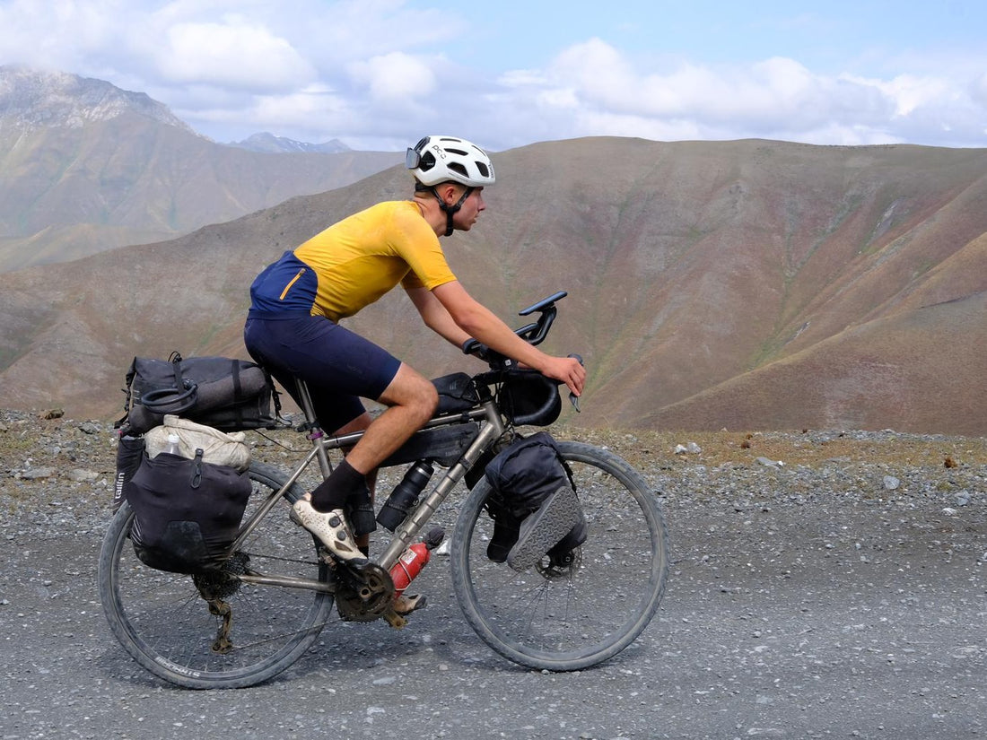 A young man rides a full laden bike across a gravel surface.