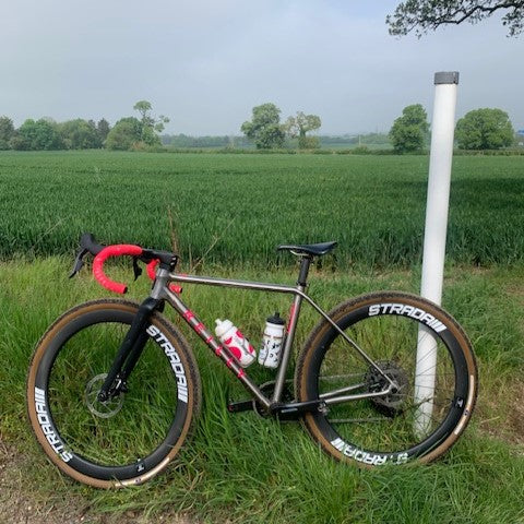 Titanium bike with pink bar tape and Reilly logo stands in a field of grass.  