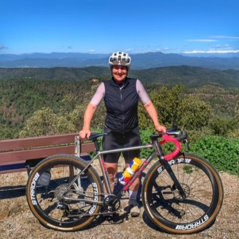 Female cyclist in front of blue sky and green landscaped with her titanium bike with pink handlebars.