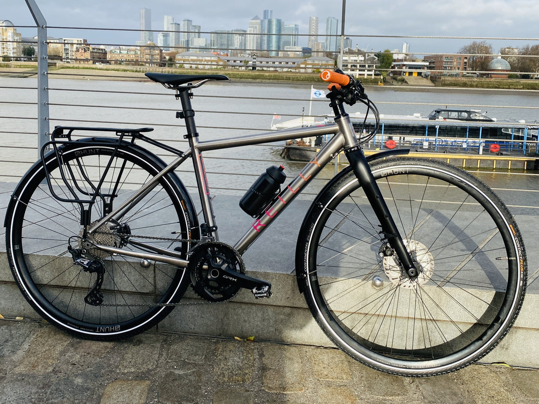 A titanium bike with a panier rack and flat handlebars in the foreground with Thames nd Canary Wharf in the background.