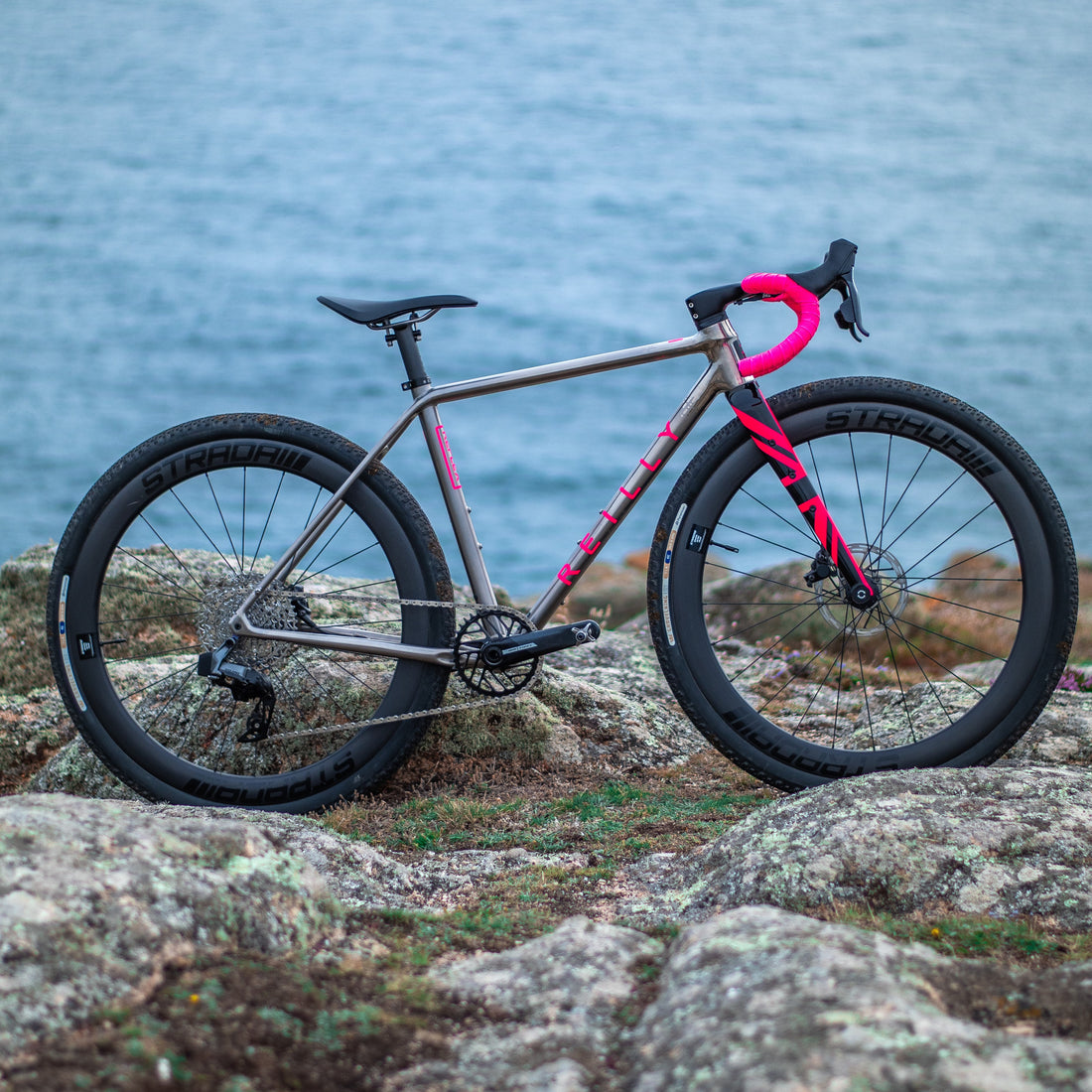 titanium bike with bright pink bar tape and pink fork pictured on rocks with the sea in the background.